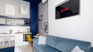 Una sala de estar moderna con un sofá azul, un comedor con una silla amarilla, una cocina con microondas y un televisor empotrado mostrando “NETFLIX”.