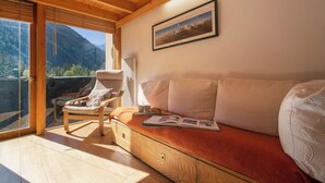 Um quarto aconchegante com teto de madeira, um sofá com almofadas, uma cadeira, uma mesa com uma xícara e um livro, e uma janela com vista para as montanhas.