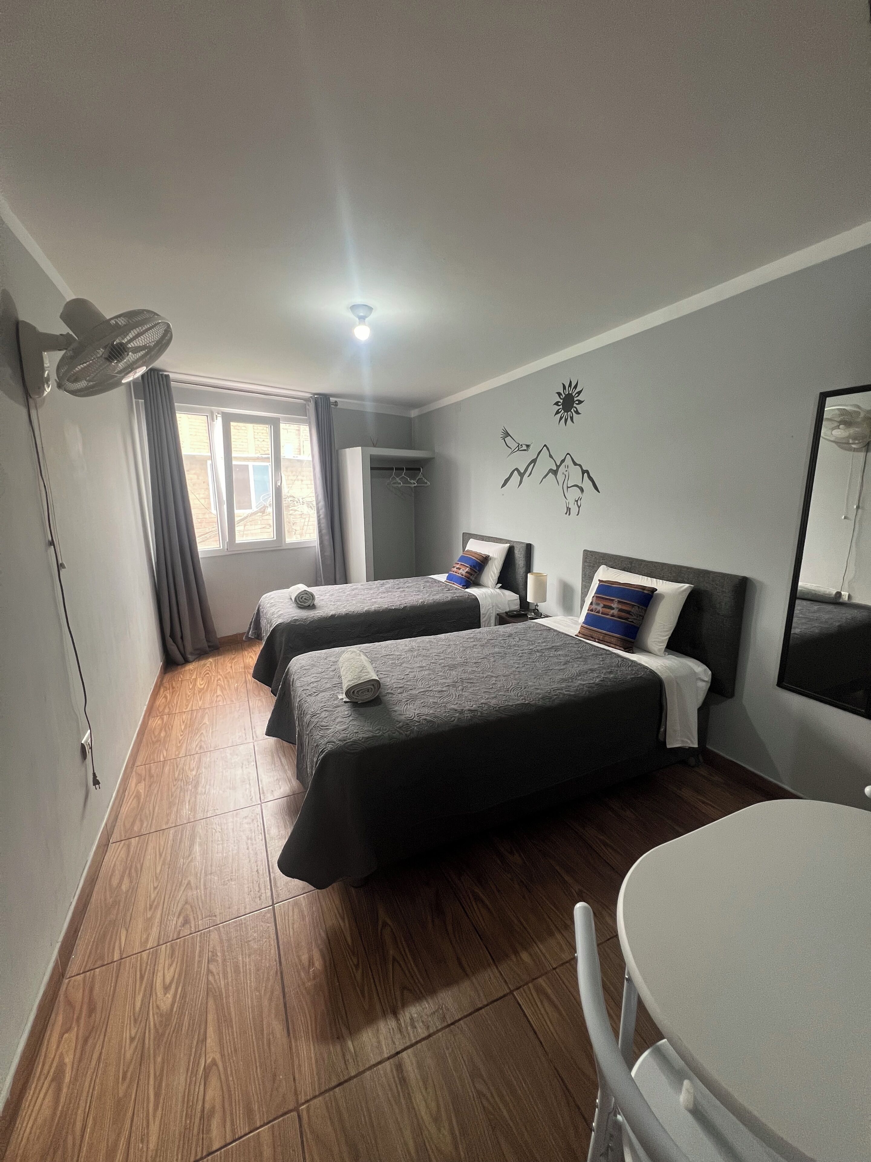 Een hotelkamer met twee bedden, een plafondventilator, een raam met gordijnen, een wandairconditioner en een ronde tafel.
