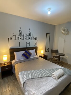 A neatly made bed with a city skyline wall art, a standing fan, and a small table with a chair in a hotel room.