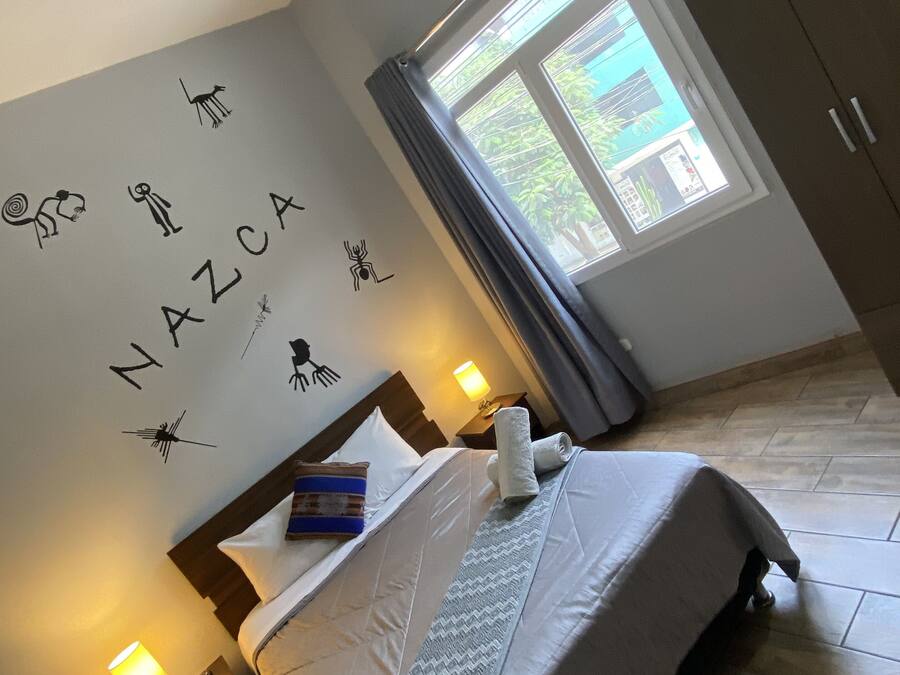 Een slaapkamer met een bed, nachtkastje, lamp, raam met gordijnen en een wand met decoratieve details.
