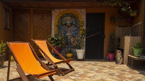 Un patio con dos sillones de madera, suelo de baldosas y un mural que representa una escena religiosa.