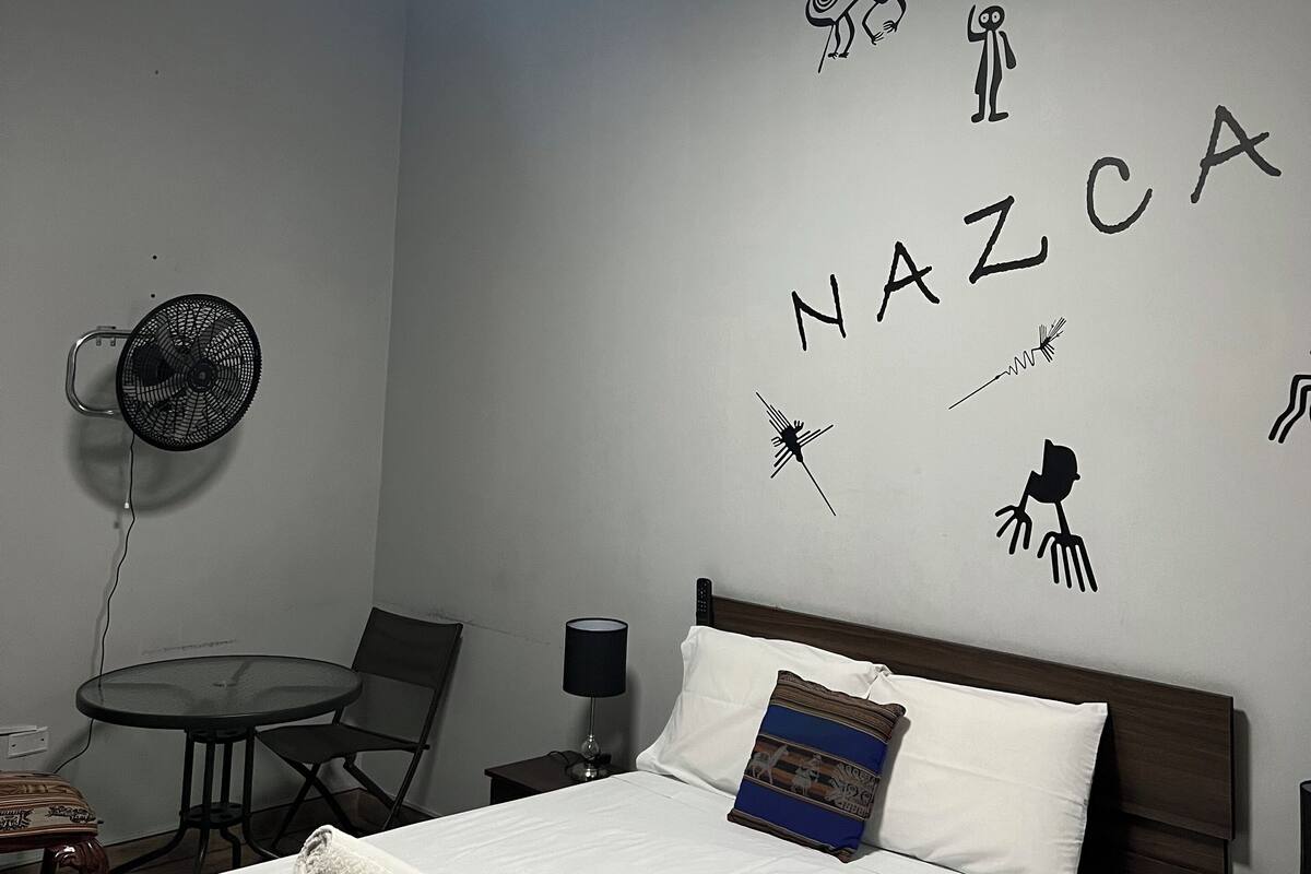 Une chambre d’hôtel avec un lit, un ventilateur fixé au mur, une table avec des chaises et un mur couvert de gribouillis.