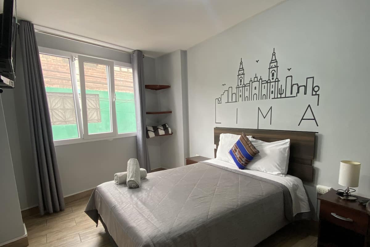 Une chambre d’hôtel avec un lit, une table de chevet, une lampe, une fenêtre avec des rideaux, un mur orné d’une fresque représentant un paysage urbain et un luminaire au plafond.
