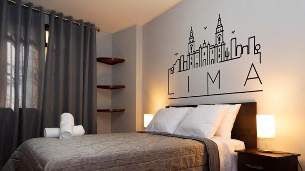 Habitación de hotel con cama, mesita de noche, lámpara, una pared con un mural de paisaje urbano y una ventana con cortinas.