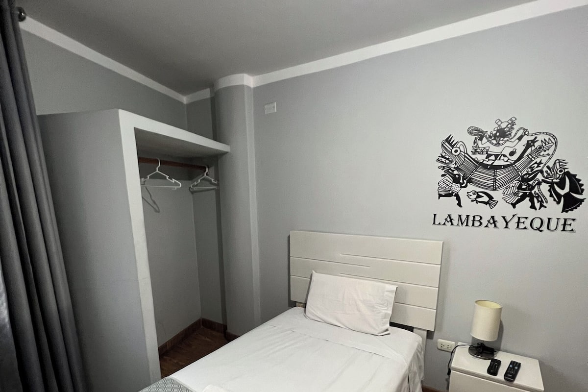 Un lit bien fait, recouvert d’une couverture à motifs, une table de chevet avec une lampe et une armoire avec des cintres.