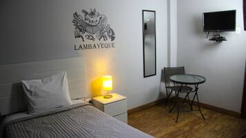 Une chambre d’hôtel comprenant un lit, une table de chevet avec une lampe, une chaise, une petite table et un téléviseur fixé au mur.