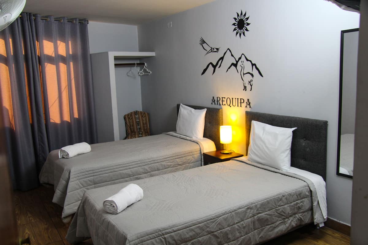 Une chambre d’hôtel avec deux lits, un ventilateur fixé au mur, une décoration murale représentant un soleil et des montagnes, et une enseigne sur laquelle on peut lire « AREQUIPA ».