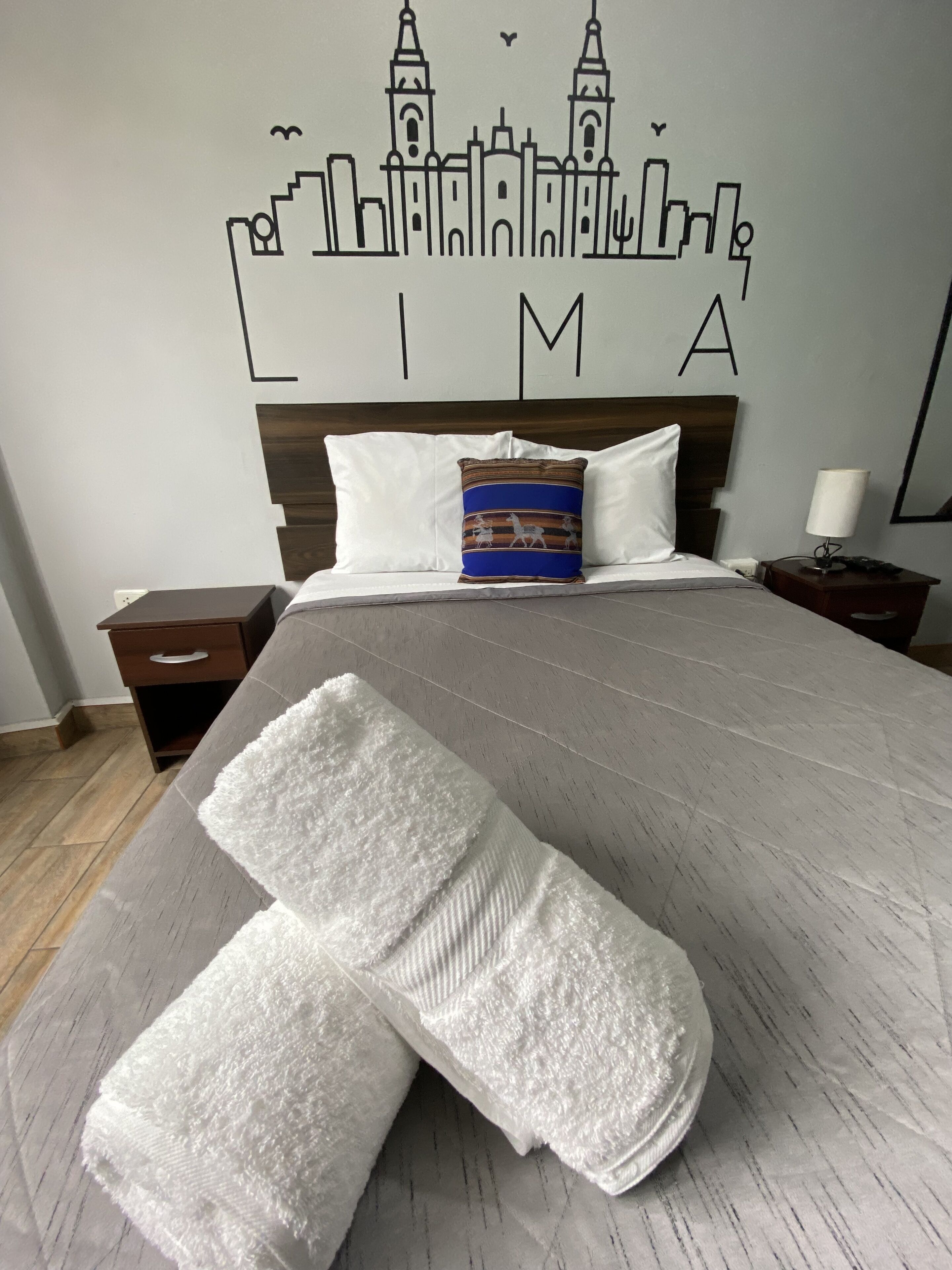 A neatly made bed with white linens, a decorative pillow, and a wall mural depicting a city skyline.