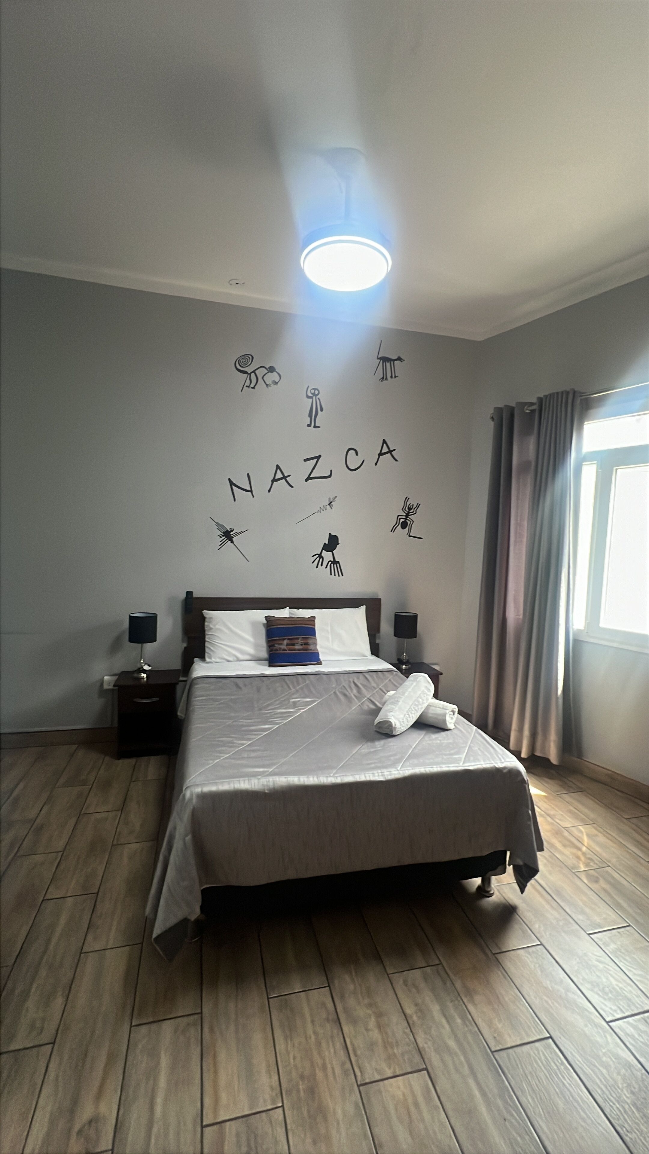 Un llit ben fet amb una almoçada i una tovallola enrotllada, una taula de nit amb una làmpada, una paret amb siluetes d'animals i la paraula "NAZCA", i una finestra amb cortines.