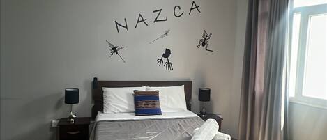 Un letto rifatto con cuscino e asciugamano arrotolato, un comodino con lampada, una parete con sagome di animali e la scritta "NAZCA", e una finestra con tende.