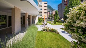 A modern building with a curved facade, a central sculpture, and a well-maintained garden with white flowers. - Prestige du Lac 27 with Balcony and Parking, Lugano, Switzerland (Lugano)