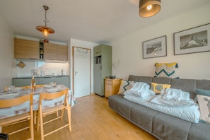 Una sala moderna con un sofá gris, una mesa de comedor de madera y sillas. Hay una zona de cocina con armarios de madera y una lámpara colgante.
