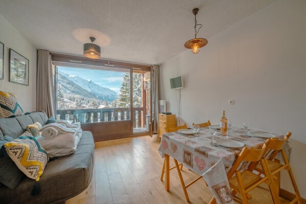 Uma sala de estar acolhedora com um sofá, uma mesa de jantar posta para dois e uma grande janela com vista para as montanhas.