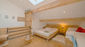 Quarto com claraboia, vigas de madeira, cama com roupa de cama branca, mesa de cabeceira com abajur e uma pequena estante com objetos decorativos.
