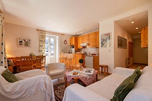 A cozy living room with two sofas, a dining table with chairs, a kitchen area with wooden cabinets, and a large window with curtains.