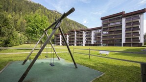 A playground with swings and a bench, surrounded by a grassy area and a multi-story building with balconies. - Résidence Grands Montets 110 Walk to Ski Lift | Argentière Chamonix, Argentière (Chamonix), France (Argentière (Chamonix))