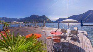 Een houten terras met buitenmeubilair, parasols en uitzicht op een meer en bergen.