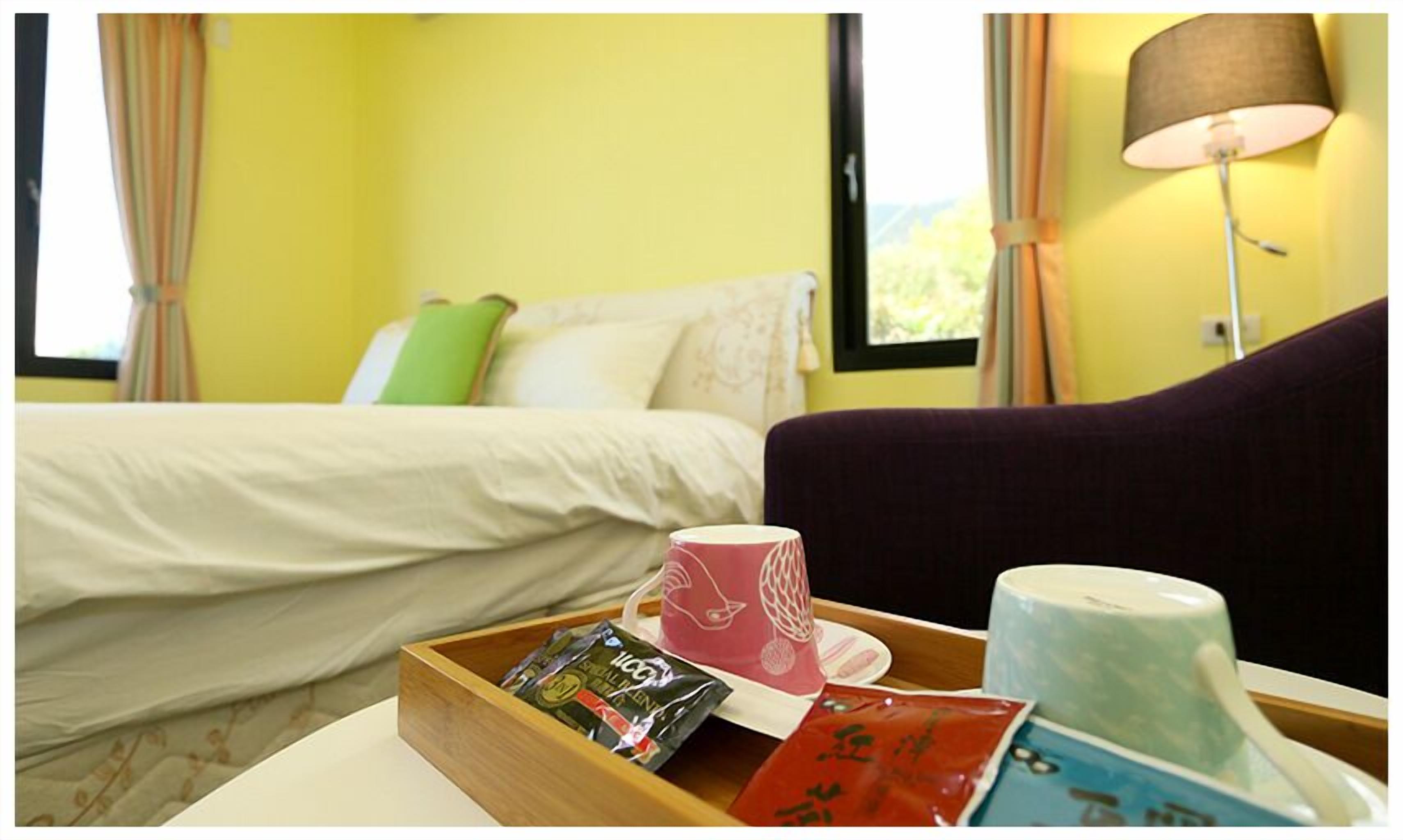 A hotel room with a bed, a bedside table with a tray of snacks, a lamp, and a view of greenery outside.