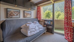 Ein Zimmer mit grauem Sofa, zwei gerahmten Bildern an der Wand, einem Holzregal und Blick auf eine bewaldete Landschaft durch ein Fenster.