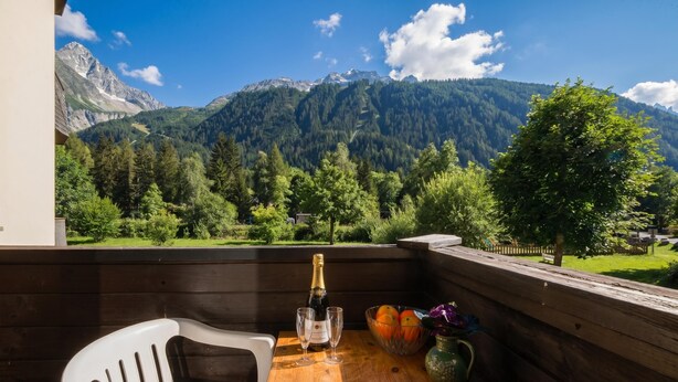 Ein Balkon mit Holztisch, zwei Gläsern, einer Flasche, einer Obstschale und einem Stuhl.