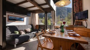 Ein gemütliches Zimmer mit Sofa, Esstisch, Stühlen, Fernseher und Blick auf die Berge.
