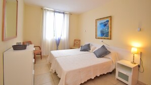 Habitación de hotel con dos camas, un escritorio con una cesta, una silla, una mesita de noche con una lámpara y un cuadro en la pared.