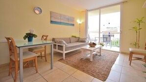 Un salón con una mesa de comedor de vidrio, un sofá y una mesa baja. Se ve una ventana grande con vistas a un edificio.