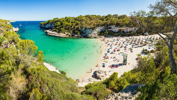 Una playa costera con aguas turquesas cristalinas, un acantilado rocoso y una zona de arena con sombrillas y tumbonas.
