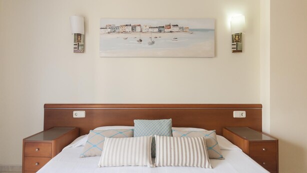 Ett hotellrum med en sänggavel i trä, en säng med randiga och geometriskt mönstrade kuddar, en tavla som föreställer ett kustlandskap och sänglampor.