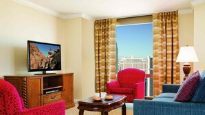 A hotel room with a television, a red armchair, a blue sofa, a wooden coffee table, and a view of a cityscape through large windows.