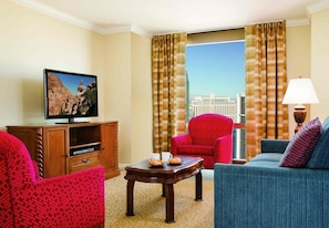 A hotel room with a television, a red armchair, a blue sofa, a wooden coffee table, and a view of a cityscape through large windows.