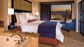 Habitación de hotel con una cama grande, un sofá, vista panorámica de la ciudad, baño con lavabo y espejo, y mesita de noche con lámpara y teléfono.