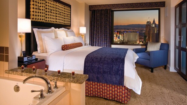 A hotel room with a large bed, a sofa, a view of the cityscape, a bathroom with a sink and mirror, and a nightstand with a lamp and phone.