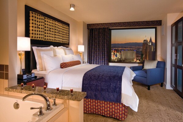 A hotel room with a large bed, a sofa, a view of the cityscape, a bathroom with a sink and mirror, and a nightstand with a lamp and phone.