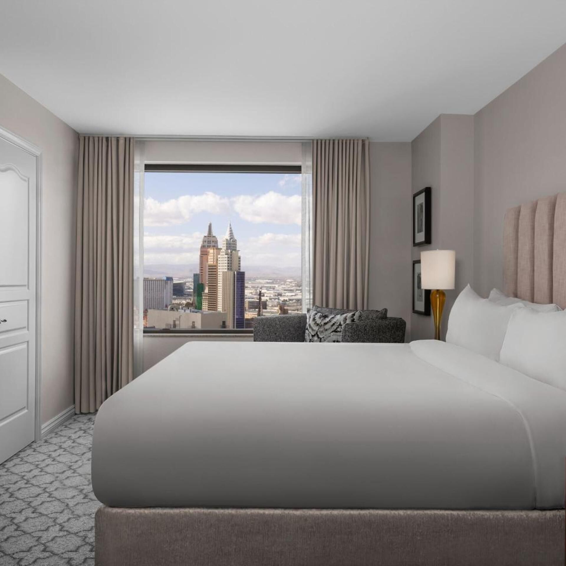 A hotel room with a large bed, a view of a cityscape, a desk with a lamp, and a chair.