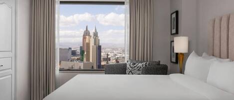 A hotel room with a large bed, a view of a cityscape, a desk with a lamp, and a chair.