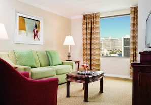A hotel room with a green sofa, a red armchair, a wooden coffee table, a lamp, a TV, and a large window with a city view. - Marriott's Grand Chateau (Las Vegas)