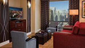 Habitación de hotel con un ventanal grande que ofrece vistas a la ciudad, un televisor de pantalla plana, un sofá rojo, un sillón gris y una mesa de centro de madera.