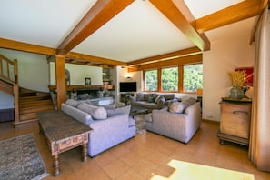 A spacious living room with a high ceiling, built-in wooden beams, a fireplace, and a large window providing natural light.