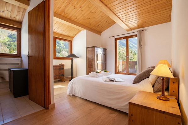 A bedroom with a wooden ceiling, a bed with a white bedspread, a nightstand with a lamp, and a view of the outdoors through large windows.
