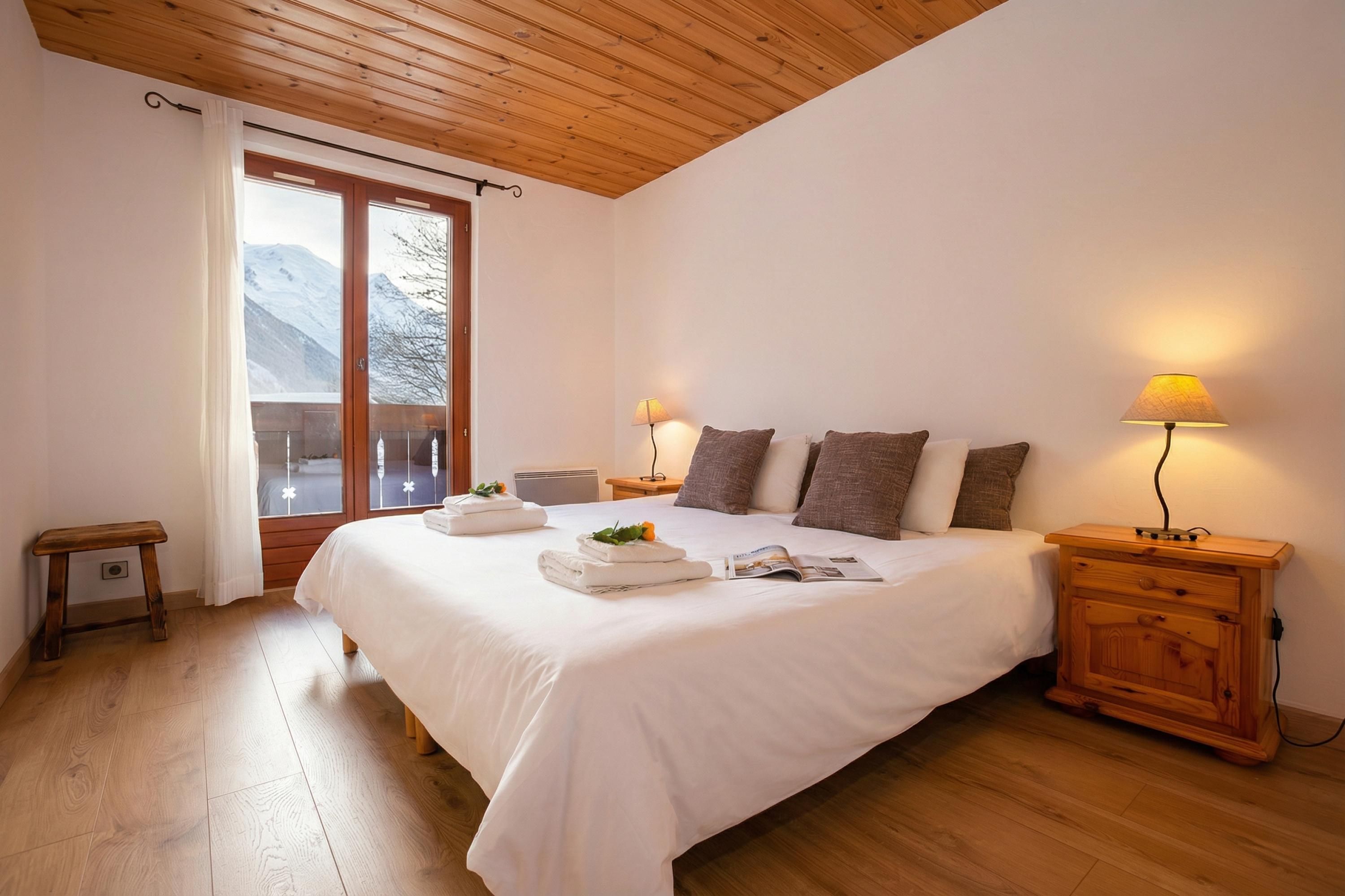 A bedroom with a large bed, two bedside tables, a wooden nightstand, a window with a view of mountains, and a wooden ceiling.