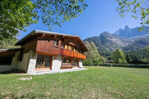 A wooden chalet with a stone foundation, large windows, and a balcony overlooking a grassy area with mountains in the background.