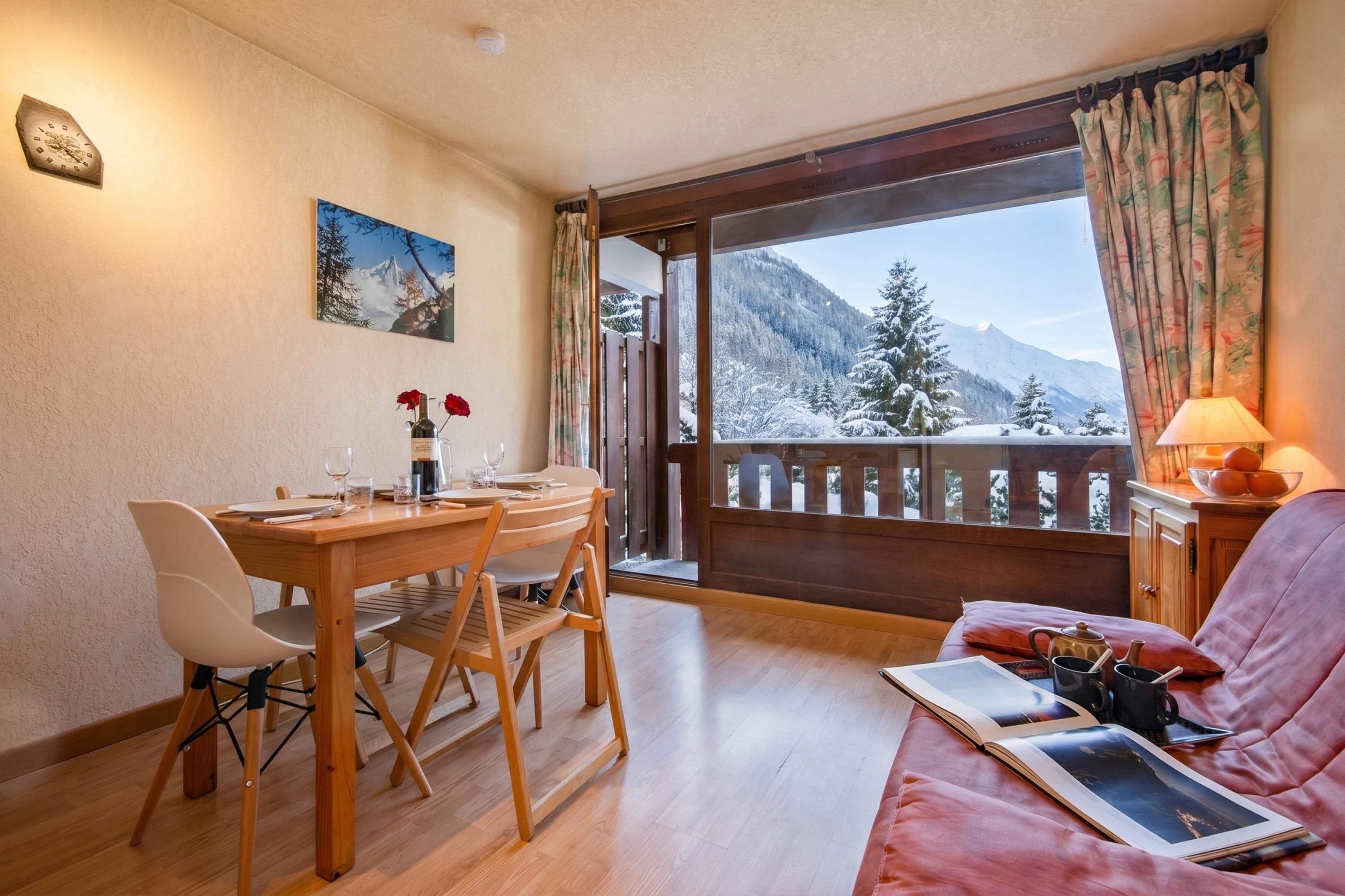 A cozy room with a wooden dining table, chairs, a sofa, a window with a view of snow-covered mountains, and a painting on the wall.