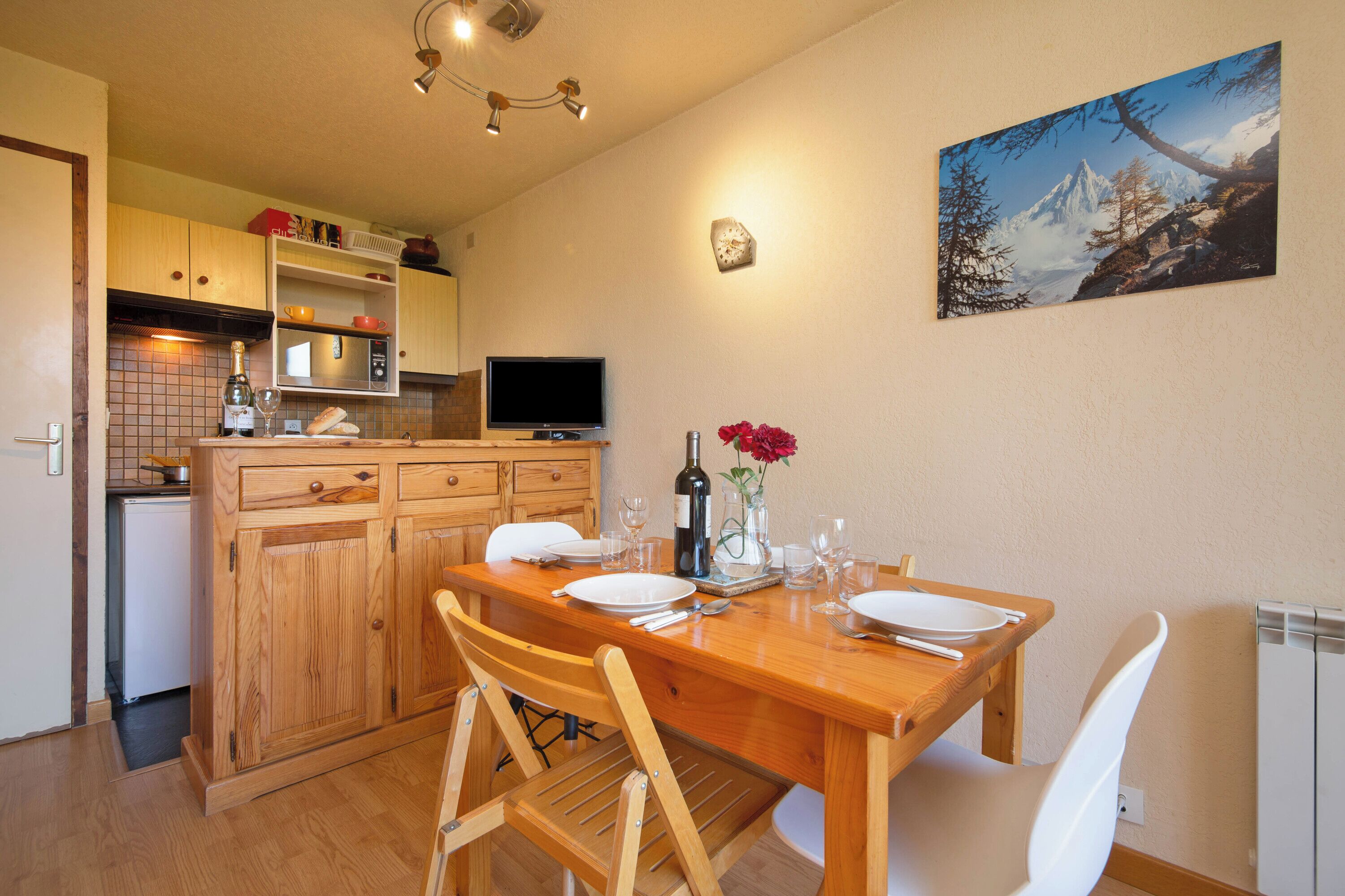A dining area with a wooden table set for two, a television in the background, and a mountain landscape painting on the wall.