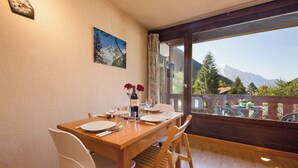Un comedor con una mesa de madera, sillas blancas, un cuadro y vistas a montañas a través de amplios ventanales.
