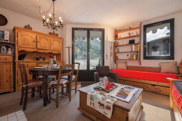 A cozy room with a dining table, chairs, a sofa, a bookshelf, and a window with a view of the outdoors.