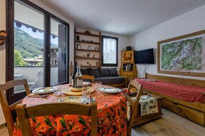 Un coin repas avec une table dressée pour deux personnes, un canapé, un téléviseur et une grande fenêtre donnant sur les montagnes.