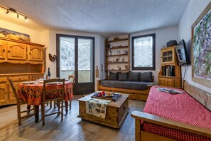 A cozy living room with a wooden dining table, chairs, a sofa, and a coffee table. There are two windows with curtains, a bookshelf, and a television on a wooden stand.