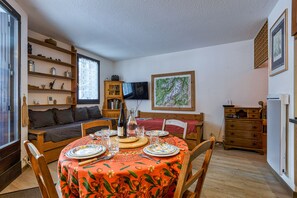 Un coin repas accueillant, avec une table ronde dressée pour quatre personnes, entourée de chaises en bois. Un téléviseur est fixé au mur, et une bibliothèque est visible sur la gauche.